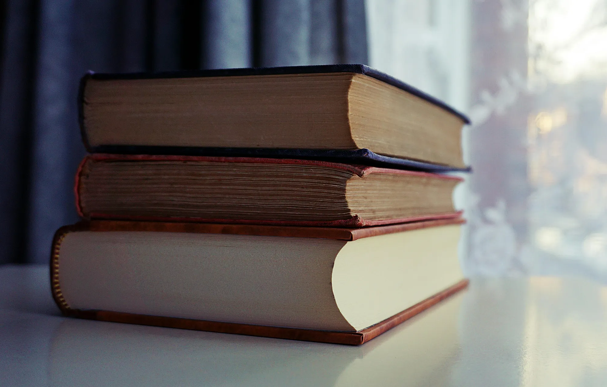 A stack of old books sits on a shelf by a sunlit window, capturing a sense of quiet study.