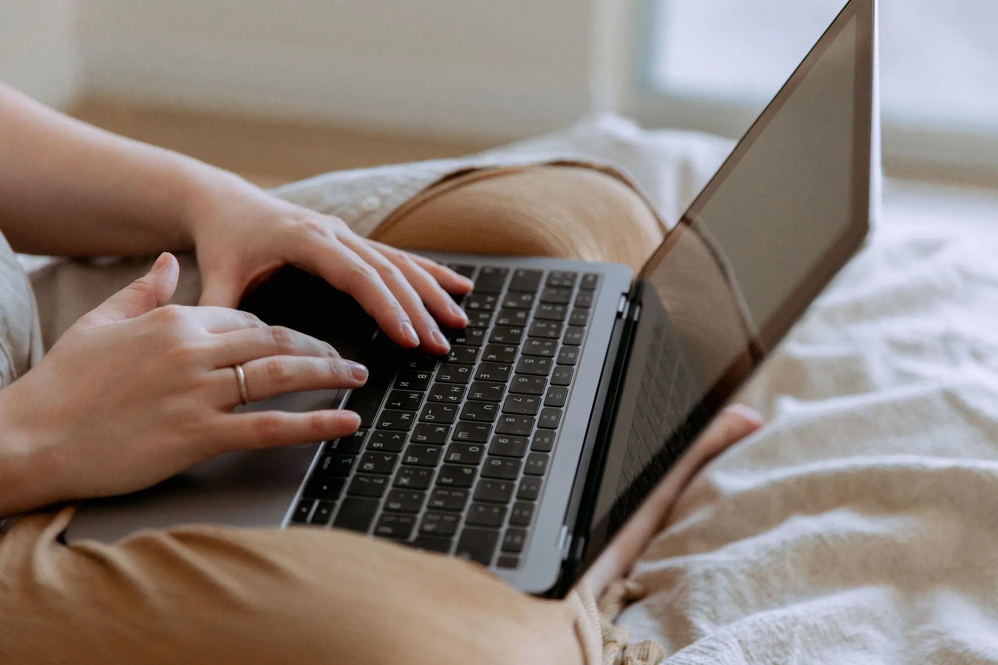 Crop anonymous female in casual wear sitting on bed with legs crossed holding laptop on knees and typing while working at home